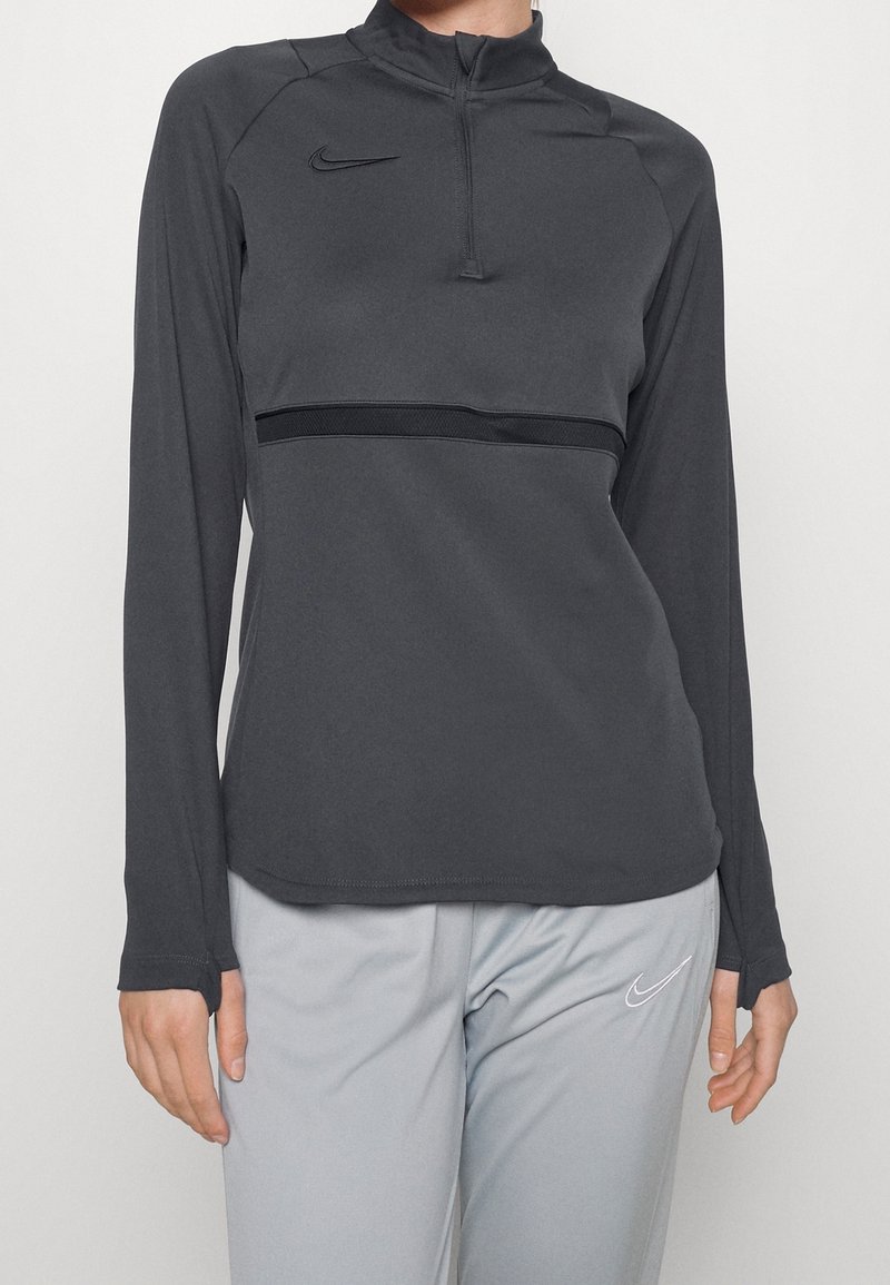 Personne portant un haut de sport gris foncé à manches longues avec une fermeture éclair sur un quart et un pantalon gris clair, tous deux avec des logos Nike.