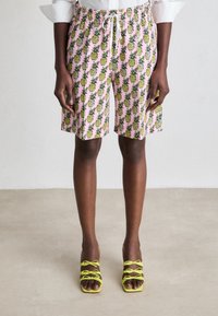 Henkilö, jolla on päällään vaaleanpunaiset polvipituiset shortsit, joissa on keltaisia ananaksia, ja limenvihreät naytööllä varustetut korkokengät, seisoo neutraalilla lattialla.