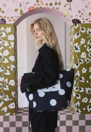 Borsa tote in denim con grandi pois bianchi, caratterizzata da un pannello di accento blu chiaro e ricami floreali, portata da un modello in un giubbotto scuro.
