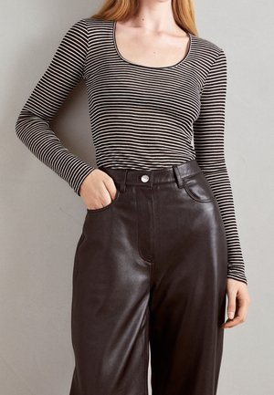Frau in einem schwarz-weiß gestreiften Langarmoberteil, das in eine hoch taillierte schwarze Lederhose gesteckt ist, mit der Hand in der Tasche vor einem schlichten Hintergrund.