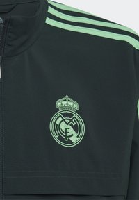 Chaqueta deportiva verde oscuro con acentos a rayas verde claro. Presenta un logo bordado con una corona y un cuello con cremallera. Textura de tela suave.
