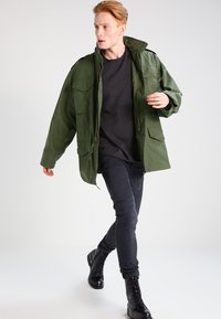 Jachetă parka de culoare verde măsliniu, cu multiple buzunare frontale, cu fermoar și închidere cu nasturi, purtată peste o cămașă neagră și pantaloni negri mulți, cu bocanci cu șiret.
