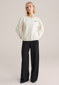 Krema pulover z rebrastimi manšetami, z enostavnim logotipom, kombiniran z visokim pasom črne široke hlače in temnimi čevlji.
