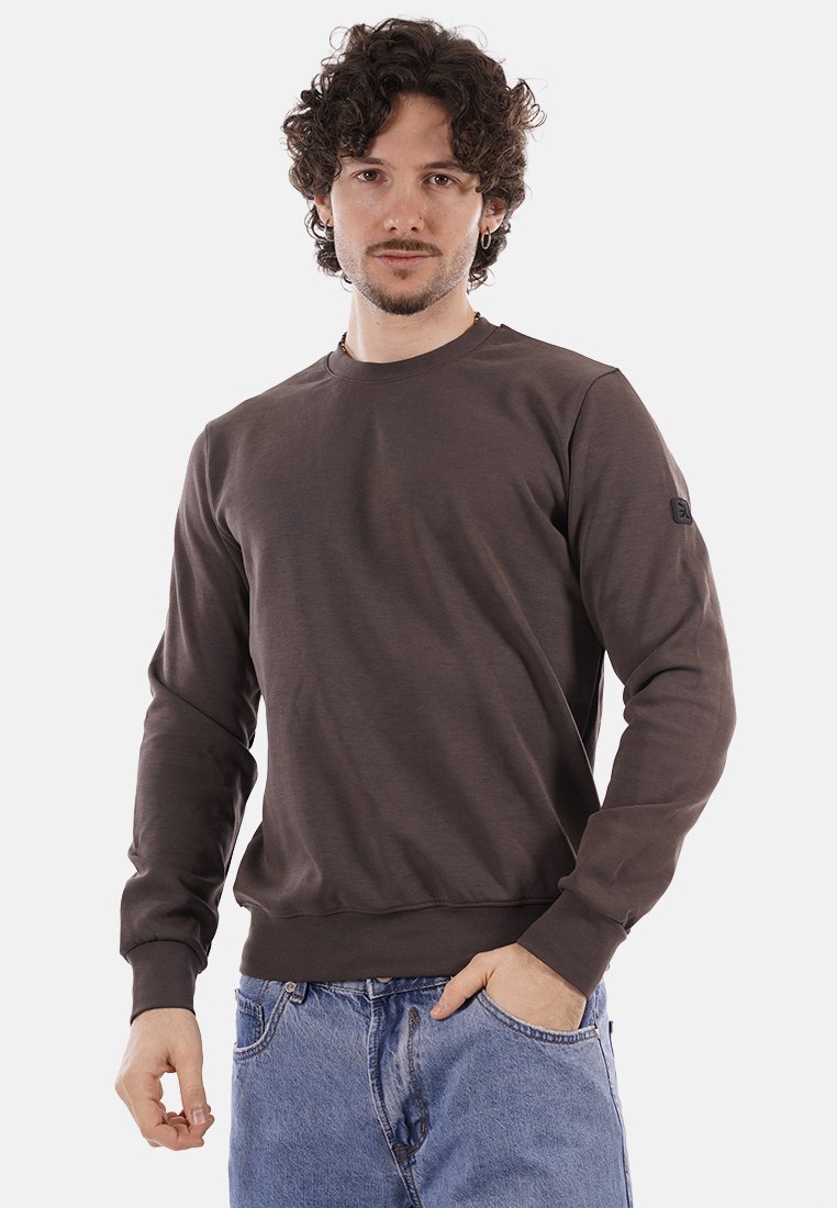 Jeune homme aux cheveux bouclés portant un sweat-shirt marron à manches longues et un jean bleu, debout avec une main dans la poche sur fond blanc.