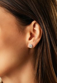 Nahaufnahme eines Frauenohres mit langen braunen Haaren, das einen runden silbernen Ohrstecker mit kleinen funkelnden Steinen trägt.