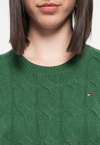 Zielony sweter z dzianiny z wzorem warkoczowym, okrągłym, ściągaczowym dekoltem i małym logo w kolorze czerwonym, białym i niebieskim po lewej stronie.