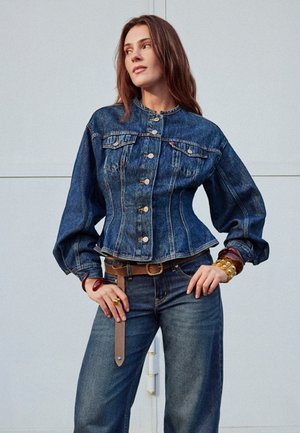 Naine kannab liibuvaid teksatagi puffvarrukatega ja kõrge vöökohaga pükse, poseerides käed puusal ühtlase tausta ees.