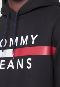 Černá mikina s kapucí s bílým textem "TOMMY" a "JEANS". Obsahuje červený horizontální pruh a bílé akcenty. Nastavitelné námořnické šňůrky na kapuci.