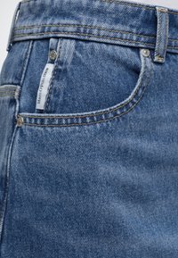 Μπλε τζιν τζιν κοντινή λήψη που δείχνει την μπροστινή τσέπη με μεταλλικά καρφιά, θηλιές για ζώνη και μια μικρή λευκή ετικέτα με την επιγραφή «Marc O'Polo Denim».