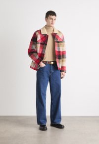 Red and beige plaid jacket with a beige shearling collar, worn over a beige turtleneck, paired with blue wide-leg jeans and black shoes.