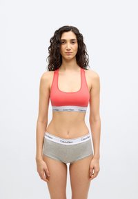 Γυναίκα με σγουρά μαλλιά που φοράει κοραλλί αθλητικό μπουστάκι και γκρι εσώρουχα Calvin Klein, στέκεται μπροστά σε απλό λευκό φόντο.