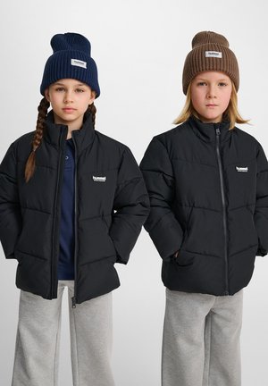 Zwei Kinder tragen schwarze Steppjacken mit hohem Kragen und Logo, kombiniert mit Strickmützen – eine in Marineblau, die andere in Braun – und grauen Sweatpants.