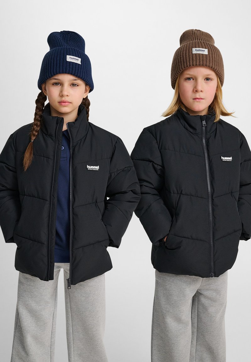Två barn bär svarta puffjackor med hög krage och logotyp, ihop med stickade mössor – en marinblå och en brun – samt grå sweatpants.