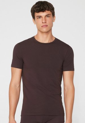 Junger Mann mit kurzen dunklen Haaren, der ein eng anliegendes dunkelbraunes T-Shirt mit kurzen Ärmeln vor einem schlichten hellen Hintergrund trägt.