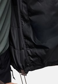 Interno di giacca a vento nera con fodera liscia e lucida; presenta un cordoncino regolabile e una texture trapuntata sul lato sinistro.