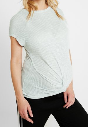 Frau trägt ein hellgraues, geripptes Kurzarmshirt mit einem Drehdetail vorne und schwarze Hosen mit Seitenstreifen.