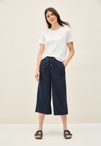 Witte katoenen t-shirt met roze logo, gecombineerd met marineblauwe wijde broek met een elastische taille en zijzakken. Zwarte sandalen.