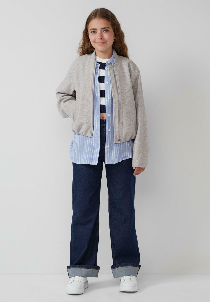 Veste bomber grise sur une chemise à rayures bleu et blanc, associée à un jean large noir et des baskets blanches. Tenue décontractée et superposée.