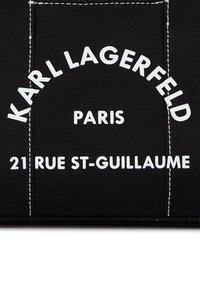 Material de lienzo negro con texto en blanco: "KARL LAGERFELD," "PARÍS," y "21 RUE ST-GUILLAUME." El texto es en negrita y tiene un borde cosido.
