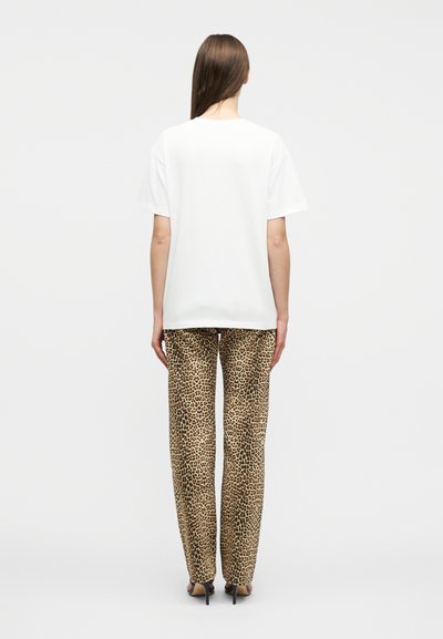 Γυναίκα που φοράει απλό λευκό T-shirt και παντελόνι με φαρδιά μπατζάκια σε λεοπάρ σχέδιο, όρθια με την πλάτη προς τα εμάς, μπροστά σε λευκό φόντο.