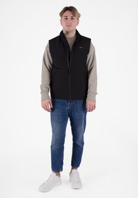 Gilet nero con chiusura a zip e tasche laterali indossato sopra una camicia beige a maniche lunghe, abbinato a jeans azzurri chiari e sneaker bianche.