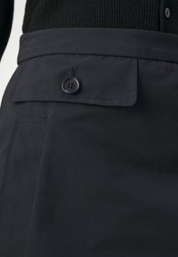 Gros plan sur la taille d'une jupe noire avec une poche à rabat sécurisée par un seul bouton noir et un pull côtelé noir rentré.