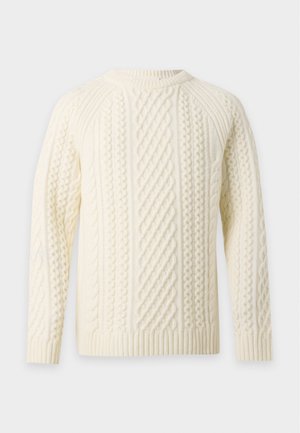 Homme debout portant un pull torsadé crème, un jean bleu ample et des baskets rouges sur un fond blanc uni.