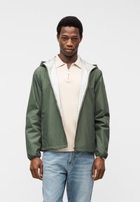 Άνδρας που φοράει ανοιχτό μπεζ πόλο με φερμουάρ, πράσινο ανοιχτό μπουφάν με κουκούλα και ανοιχτό γαλάζιο τζιν, στέκεται σε απλό φόντο.