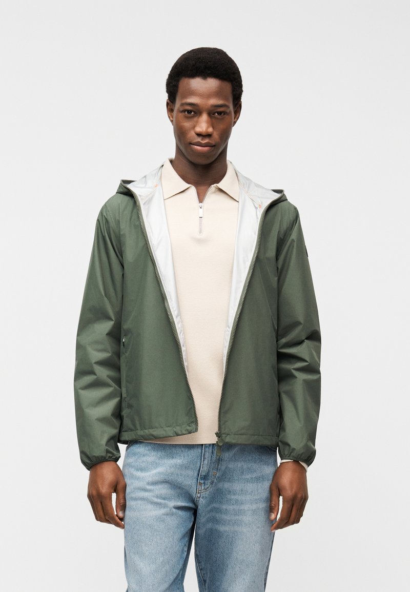 Άνδρας που φοράει ανοιχτό μπεζ πόλο με φερμουάρ, πράσινο ανοιχτό μπουφάν με κουκούλα και ανοιχτό γαλάζιο τζιν, στέκεται σε απλό φόντο.