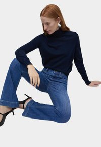 Granatowy sweter z golfem w połączeniu z niebieskimi dzwonami. Gładki materiał, dopasowany fason i czarne szpilki. Modelka pozowała w pozycji siedzącej.