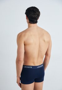 Marineblaue Baumwoll-Boxershorts mit einem weißen Bund, der das "Lacoste"-Logo trägt, von hinten betrachtet. Glatte Textur und eng anliegendes Design.