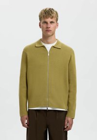 Maslinasto zeleni zip-up kardigan s ovratnikom, rebrastom teksturom, dugim rukavima i ravnim krojem. Nosite ga preko bijele majice i smešnih hlača.