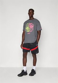 Šedé tričko s krátkým rukávem má barevnou grafiku lva, doplněné černými šortkami s červenými pruhy. Kombinováno s černými teniskami a bílými ponožkami.