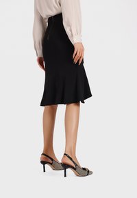 Gonna nera aderente con peplo svasato, abbinata a scarpe slingback con un motivo geometrico e un tacco basso. Blusa di colore chiaro.