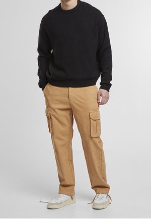 Man draagt een zwarte trui, beige corduroy cargobroek met grote zijzakken en witte sneakers met losse veters, voor een effen achtergrond.