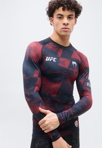 Νέος άνδρας με εφαρμοστό αθλητικό μπλουζάκι UFC με μακριά μανίκια σε μαύρο και κόκκινο χρώμα, στέκεται με τα χέρια σταυρωμένα μπροστά σε απλό ανοιχτό φόντο.