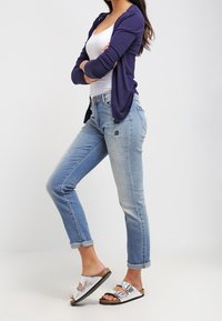 Laivastonsininen neuletakki valkoisen hihattoman paidan päällä, vaaleansiniset farkut, joissa on brodeeratut yksityiskohdat, sekä valkoiset sandaalit hopeisilla yksityiskohdilla.