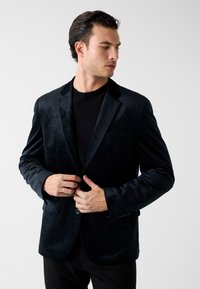 Zwarte veloursblazer met een gestructureerde afwerking, sluiting met één knoop en revers met inkeping, gecombineerd met een aansluitend zwart shirt eronder.