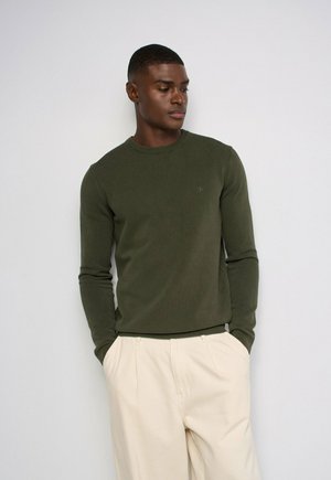 Hombre con un suéter de manga larga de color verde oliva y pantalones color crema, de pie con las manos en los bolsillos contra un fondo sencillo.