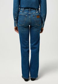 Persona vestida con jeans rectos azules y una camisa de mezclilla a juego, de pie de espaldas sobre un fondo claro y liso.