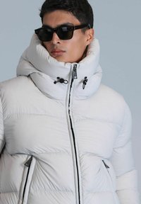 Doudoune blanche avec col montant, dotée d'une fermeture éclair à l'avant et de deux poches zippées. Texture lisse et design matelassé.