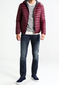 Doudoune bordeaux avec capuche grise, portée sur une chemise rayée. Associée à un jean foncé et des baskets bleu marine. Texture lisse, design léger.