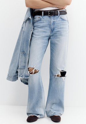 Személy világoskék, roncsolt farmernadrágot és fekete cipőt visel, miközben az egyik karján egy hozzáillő világoskék farmerdzsekit tart.