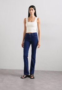 White fitted tank top, dark blue flared jeans, and black sandals. Model stands upright, medium hair, plain background.