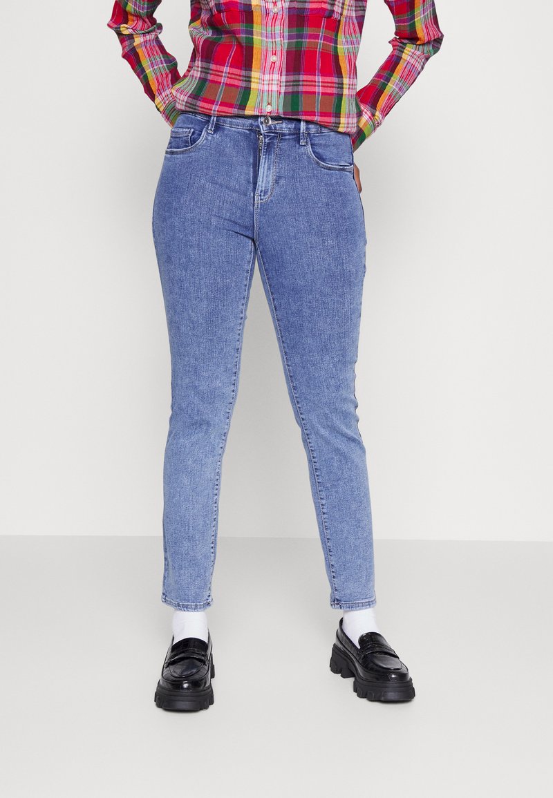 Jeans skinny azul claro com um tecido de ganga texturizado, apresentando dois bolsos frontais e uma cintura de altura média, combinados com mocassins pretos e meias brancas.