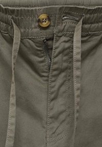 Nærbillede af kanten på khakibukser med knap, delvist åbnet lynlås og stofsnore.