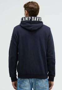 Sudadera azul marina con un gran texto blanco "CAMP DAVID" en el cuello. Presenta un color sólido, una textura suave y puños acanalados.