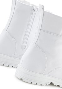 Bottines en toile blanches avec fermeture éclair et accents de lacets. La semelle est texturée pour une meilleure adhérence, présentant un design simple et utilitaire.