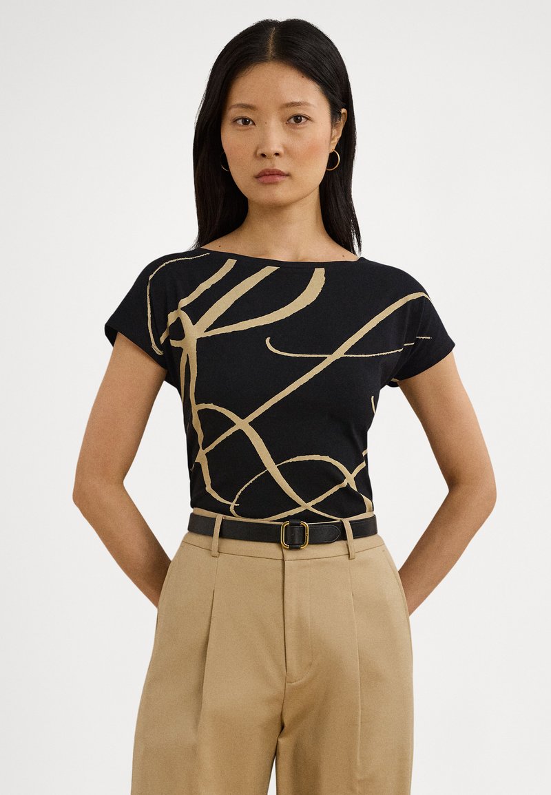 Juoda marškinėliai su auksinės abstrakčios linijos dizainu, apvalia iškirpte, trumpomis rankovėmis, derinami su smėlio spalvos aukšto liemens kelnėmis ir juodu diržu.