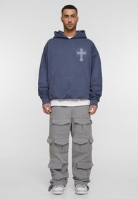 Marineblauwe hoodie met voorzak en grafisch ontwerp, gecombineerd met lichtgrijze cargobroek met meerdere zakken en een ontspannen pasvorm. Witte sneakers.